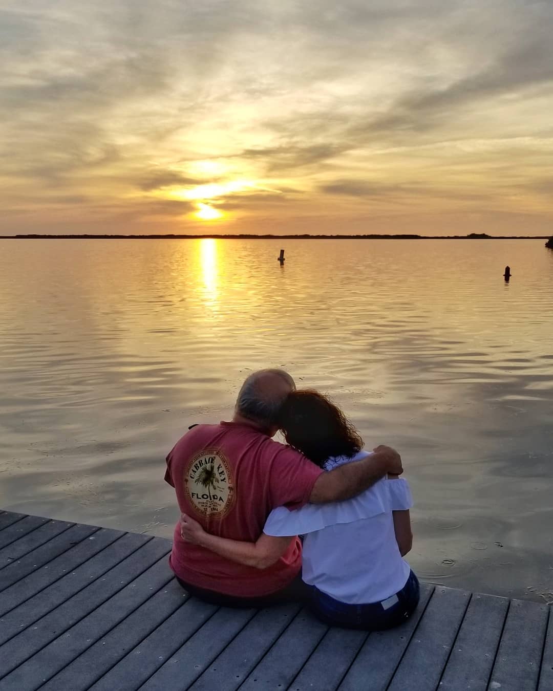 Couple sits, embraced, on the dock at Tarpon Lodge overlooking the bay and a golden sunset. 